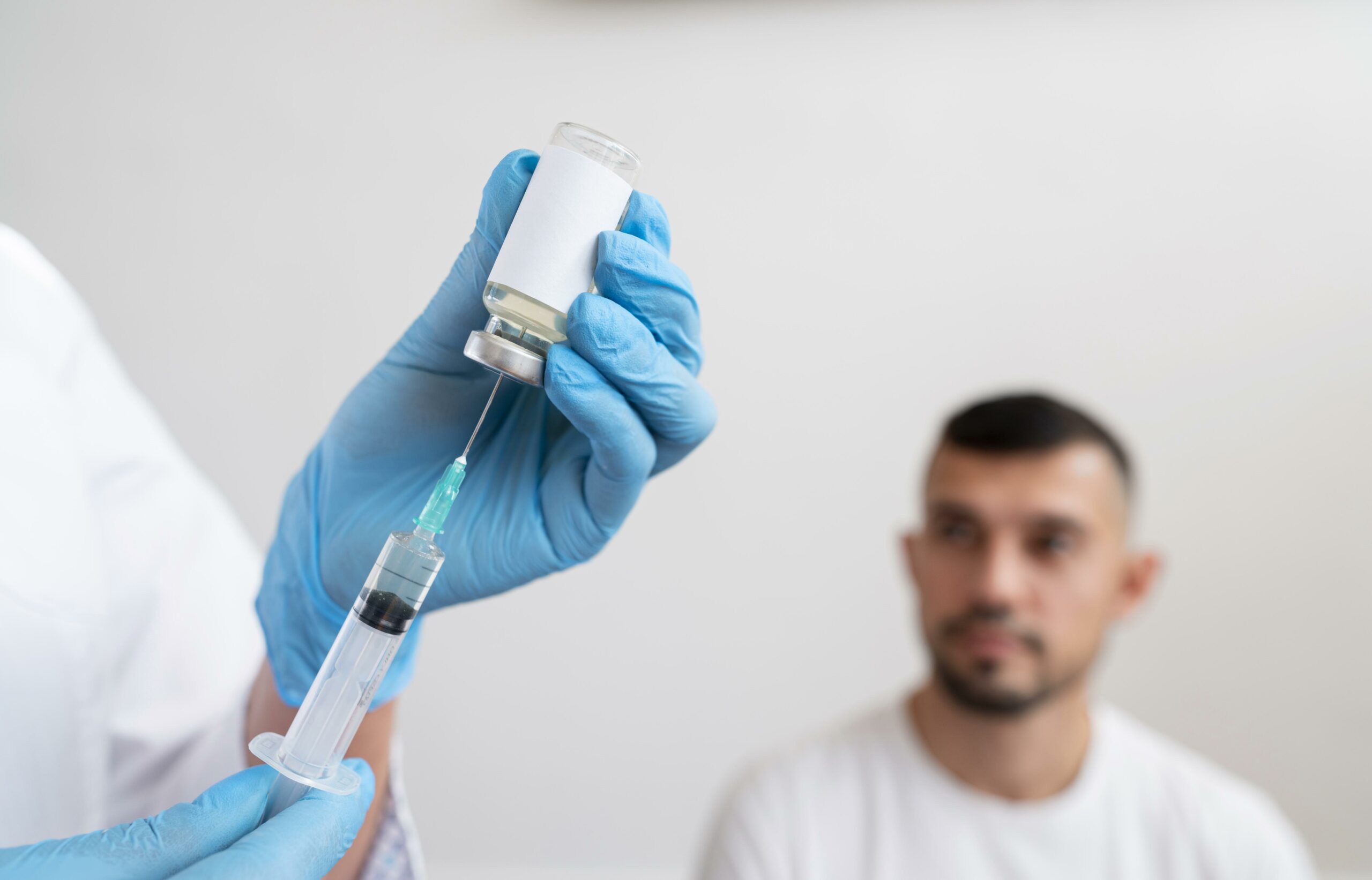 Blue-gloved hands injecting syringe into suspended vial. Slightly blurred, a dark haired bearded man in plain white tshirt sits in background looking at the blue-gloved hands injecting syringe into suspended vial