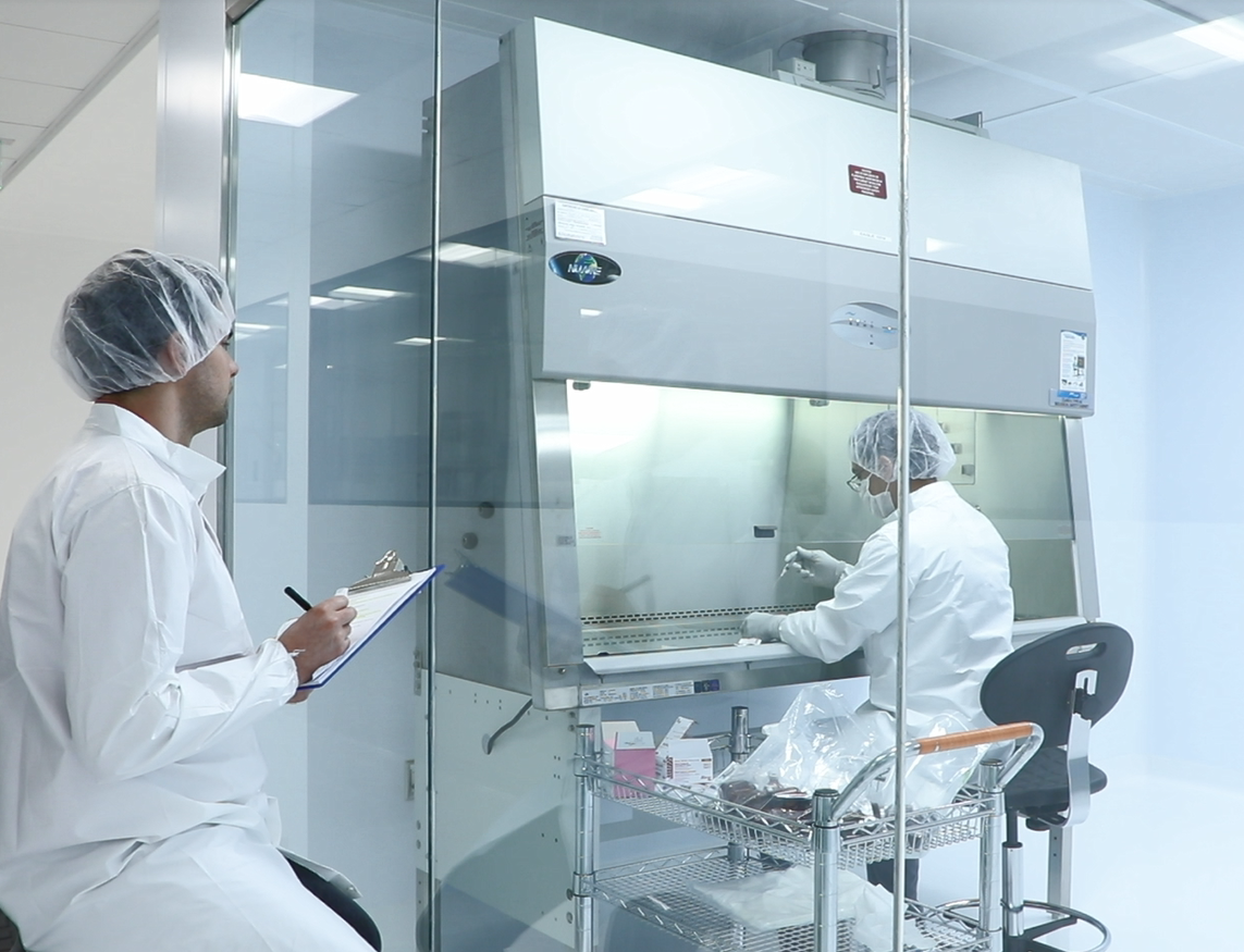 Man in lab coat and hairnet sits watching another man beyond a glass wall compounding in a sterile compounding hood.