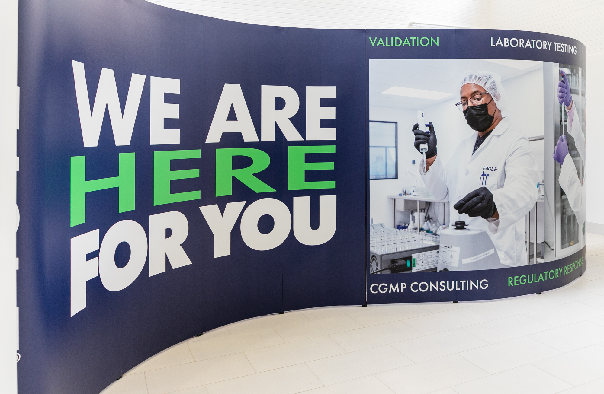 Image of Eagle's backdrop for booth for PDA. Booth shows images of two scientists performing sampling testing. The text states, "WE ARE HERE TO HELP YOU." Validation, Laboratory Testing, Training, CGMP Consulting, Regulatory Response, Remediation." On the booth's endcaps are EAGLE Science-based solutions logos.